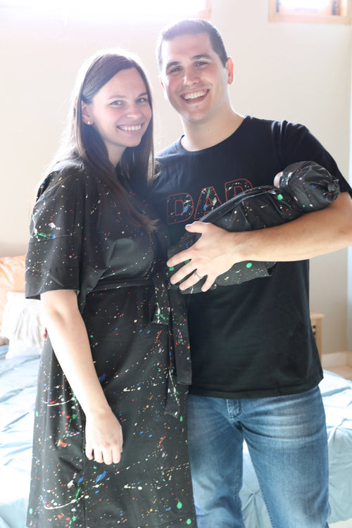 A mother and father smile while holding their newborn wrapped in a matching black paint-splatter swaddle. The mother wears a coordinating paint-splatter maternity robe, and the father wears a black “DAD” T-shirt included in the matching family hospital set. This paint splatter robe, dad shirt, and newborn swaddle bundle is ideal for maternity photos, labor and delivery outfits, hospital bag essentials, and baby announcement images.
