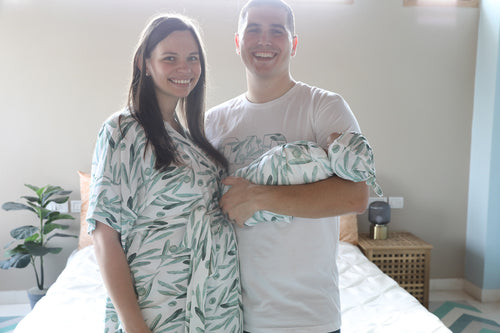 New parents holding a baby wrapped in a green and white patterned blanket in a bedroom.