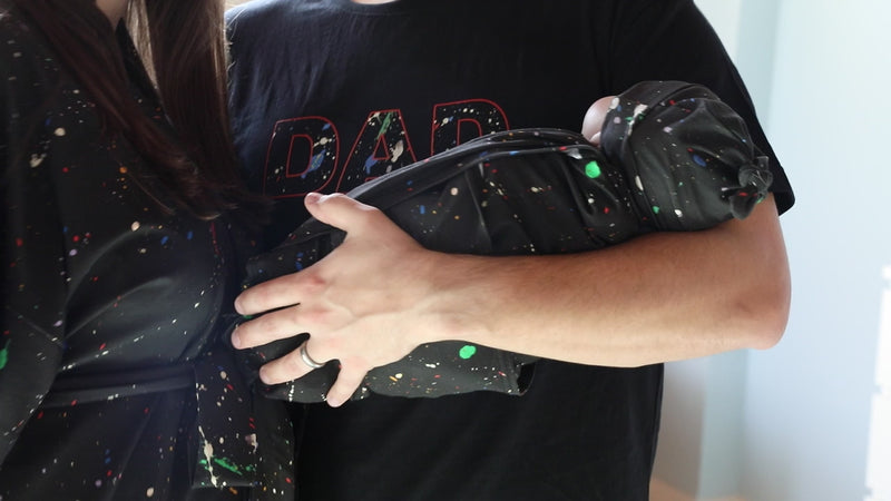 A mother and father smile while holding their newborn wrapped in a matching black paint-splatter swaddle. The mother wears a coordinating paint-splatter maternity robe, and the father wears a black “DAD” T-shirt included in the matching family hospital set. This paint splatter robe, dad shirt, and newborn swaddle bundle is ideal for maternity photos, labor and delivery outfits, hospital bag essentials, and baby announcement images.